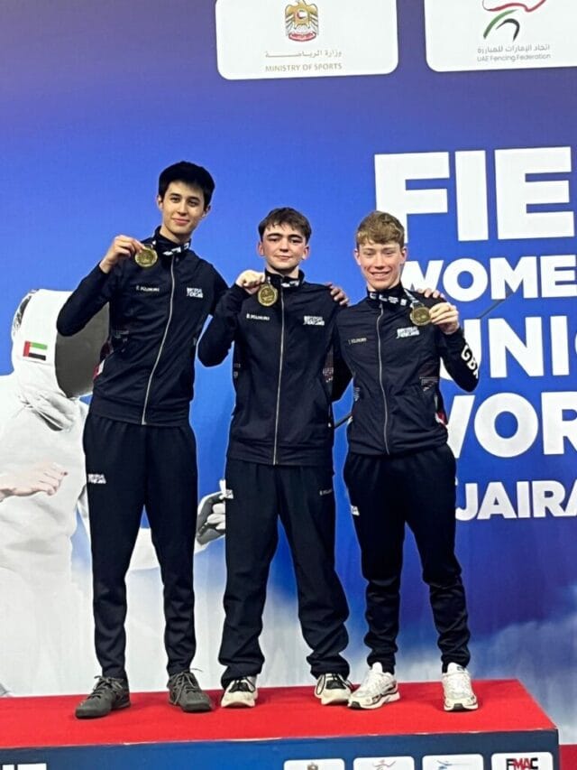 A young girl in a pink jacket and a blue scarf smiling brightly while sitting on a wooden bench in a park, surrounded by autumn trees with orange and yellow leaves.